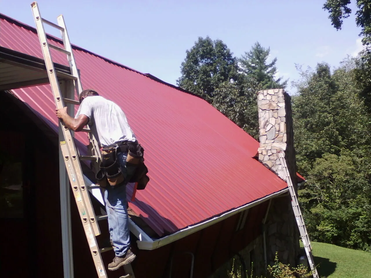 Roofer on a ladder beside a red metal roof and stone chimney — Soffit & Fascia Repair by Roofing Brothers in Drakesboro